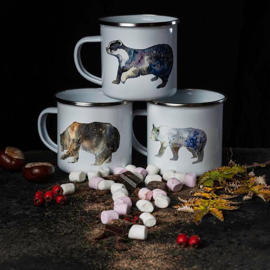 Three enamel camping mugs in a pyramid. The top mug asha picture of a badger on. The two lower mugs have illustrations of a grizzly bear and polar bear. Marshmallows, chocolate and autumn foliage surround the mugs.