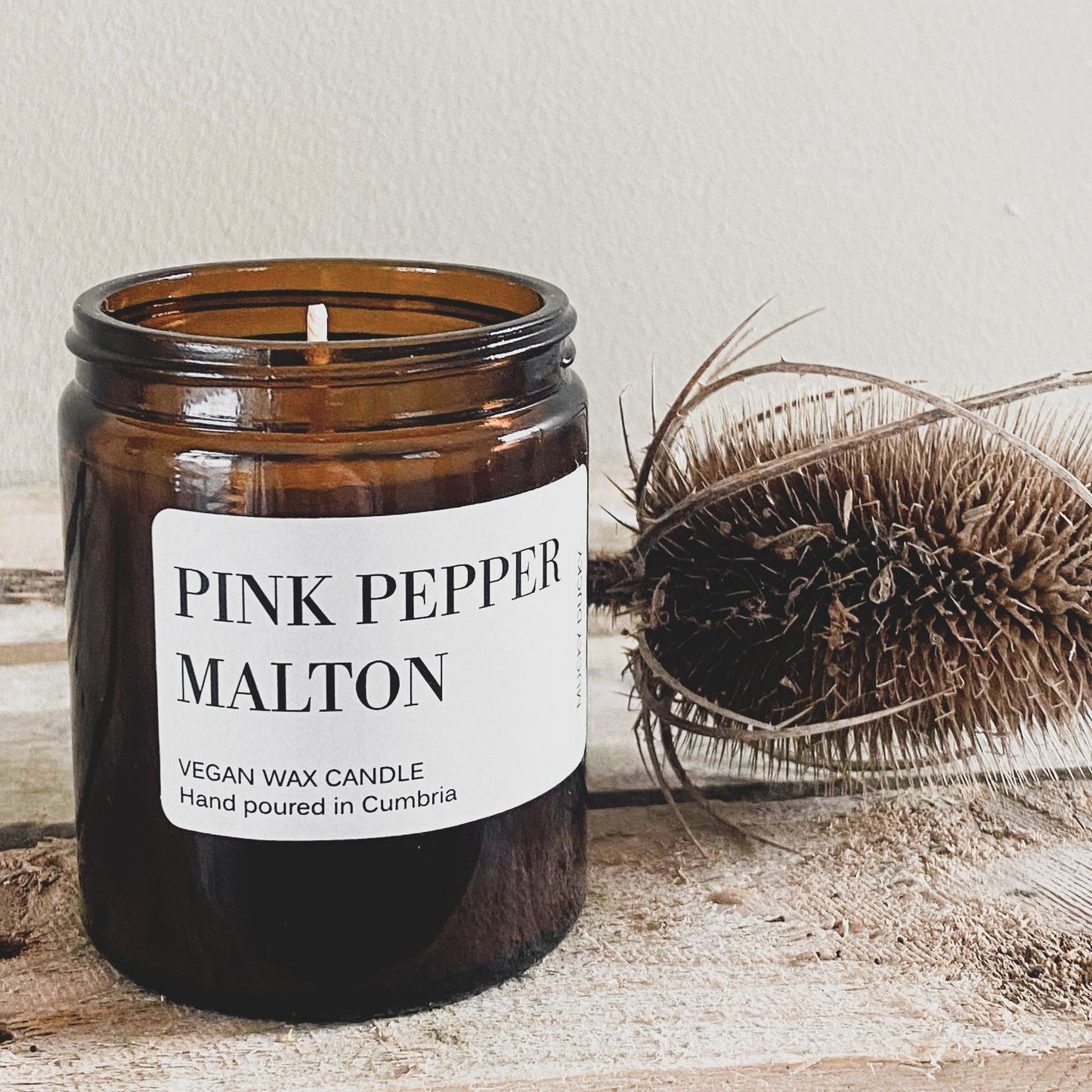Pink pepper scented candle in an amber glass jar with a dried teasel next to it
