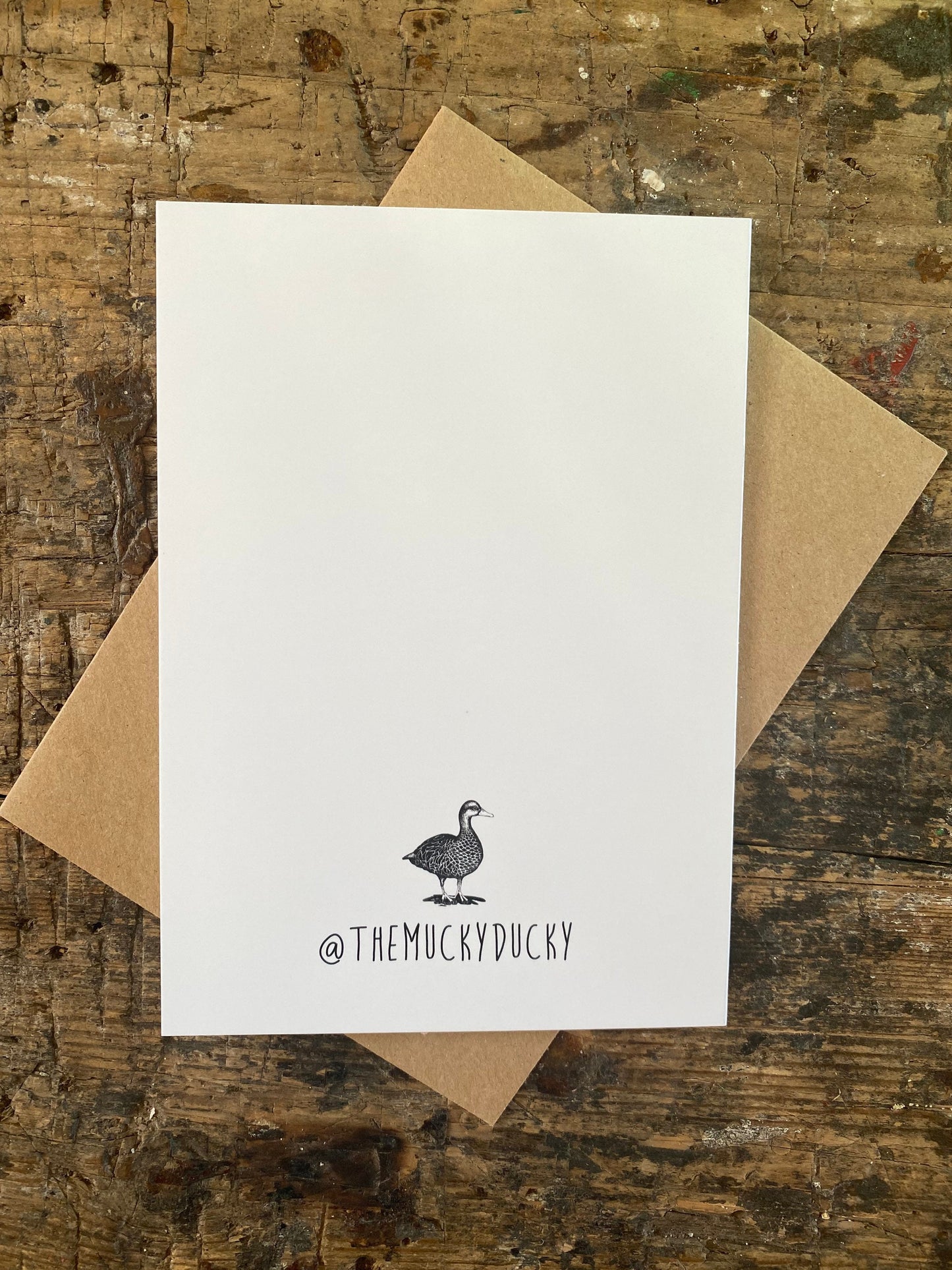 The Mucky Ducky logo on the reverse of a bear greetings card. The card has a brown envelope behind it and is placed on a rustic table.